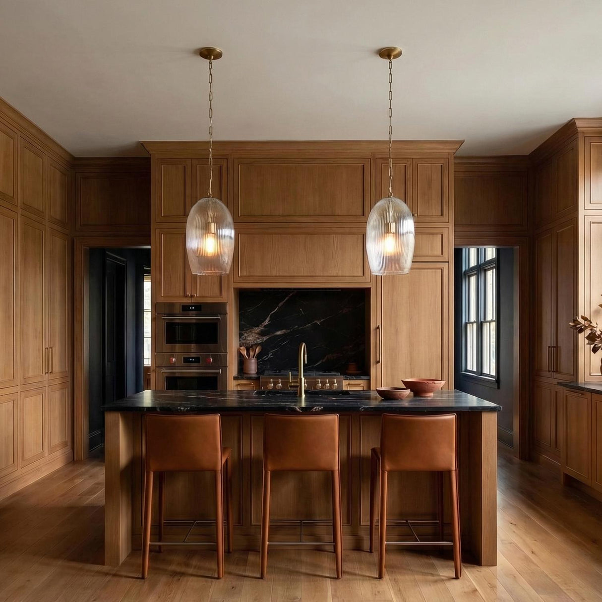A transitional kitchen features two Fiora Pendant 11", Modern Brass fixtures with ribbed glass over a black marble island surrounded by wood cabinetry and leather barstools.