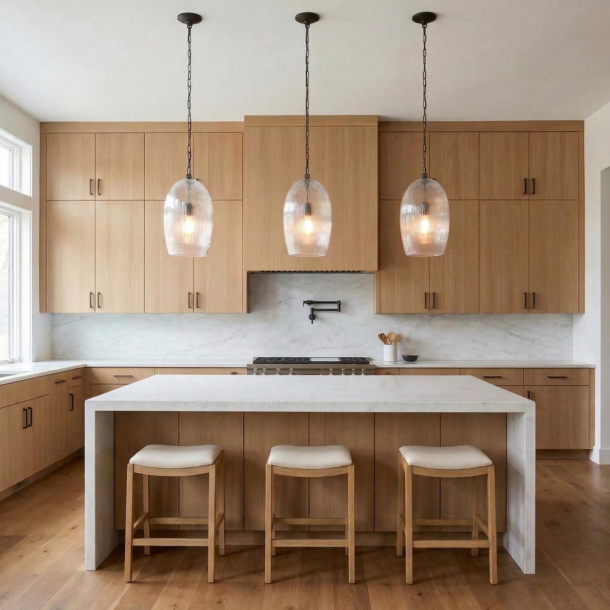 A modern kitchen featuring three Fiora Pendant 11", Aged Iron fixtures with ribbed glass over a white marble waterfall island and natural wood cabinets.