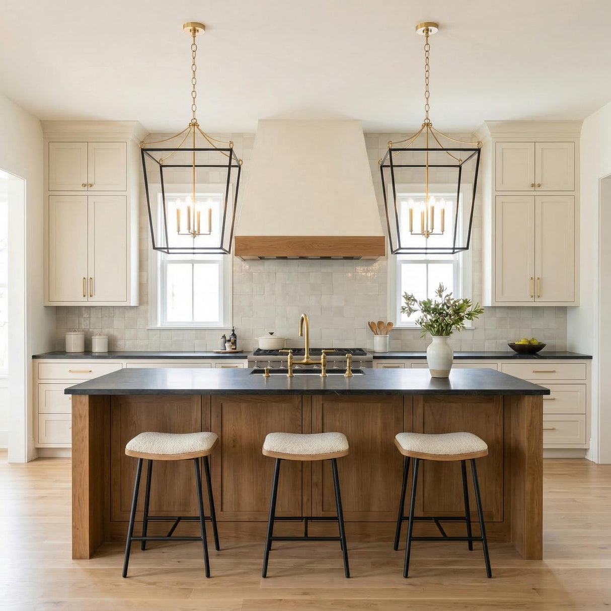 A transitional kitchen features two Cambria Extra Large Lantern Pendant 24", Matte Black and Modern Brass fixtures over a wood island with black countertops and cream cabinets.