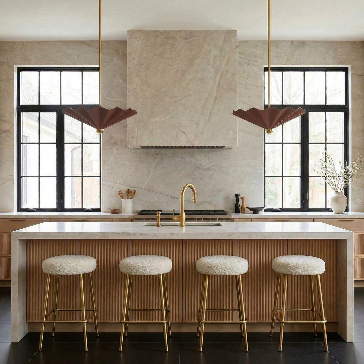 A contemporary kitchen features two Babette Large LED Pendant 25", Maroon and Brass fixtures flanking a stone range hood above a fluted wood island with four stools.