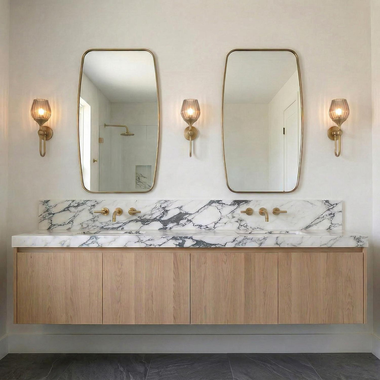 A contemporary bathroom with three aged brass and smoke taupe ribbed glass wall sconces flanking two gold framed mirrors over a marble countertop and oak vanity.