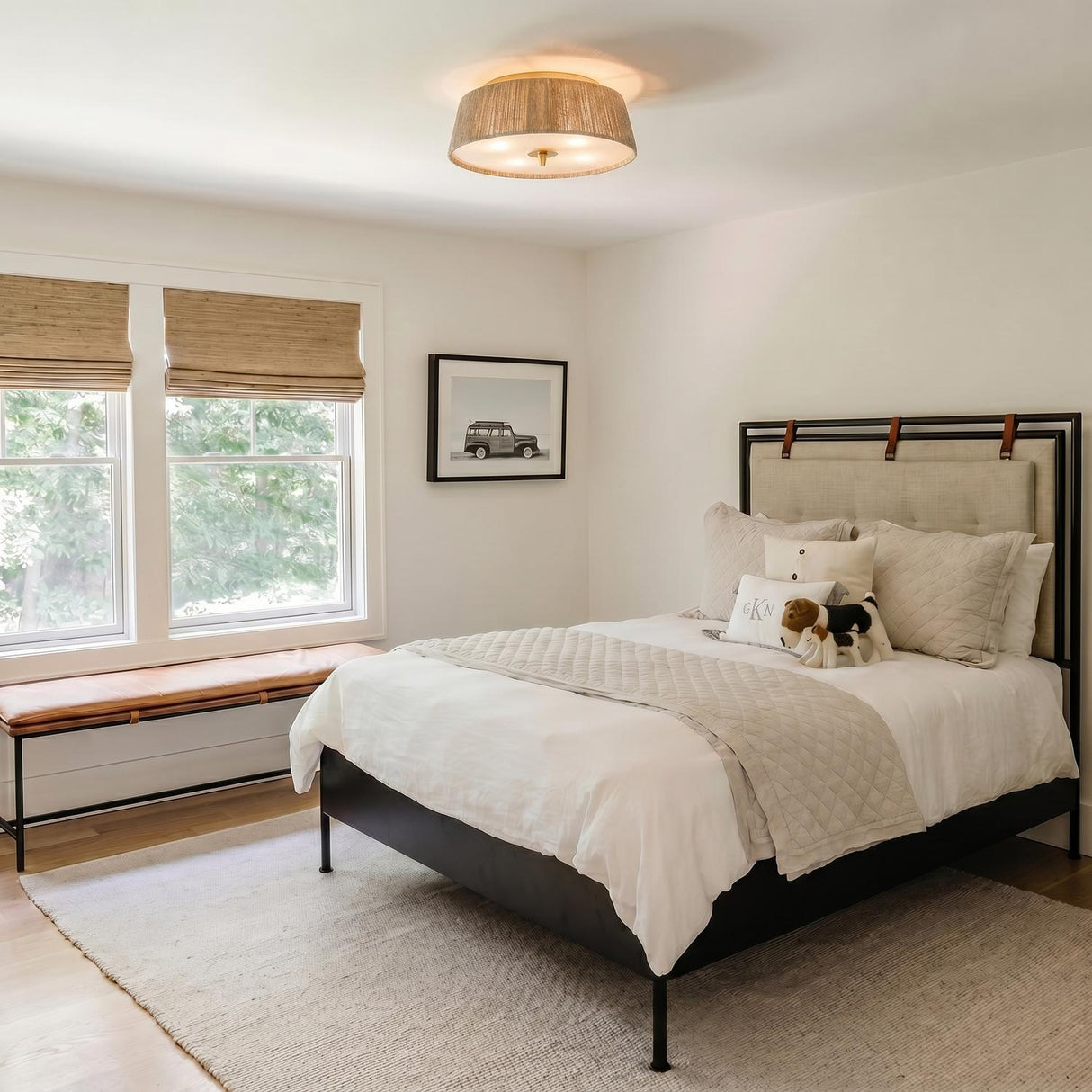 A transitional bedroom with the jute and aged brass Pascal Flush Mount 18" installed in the ceiling center above a black bed frame and leather bench.