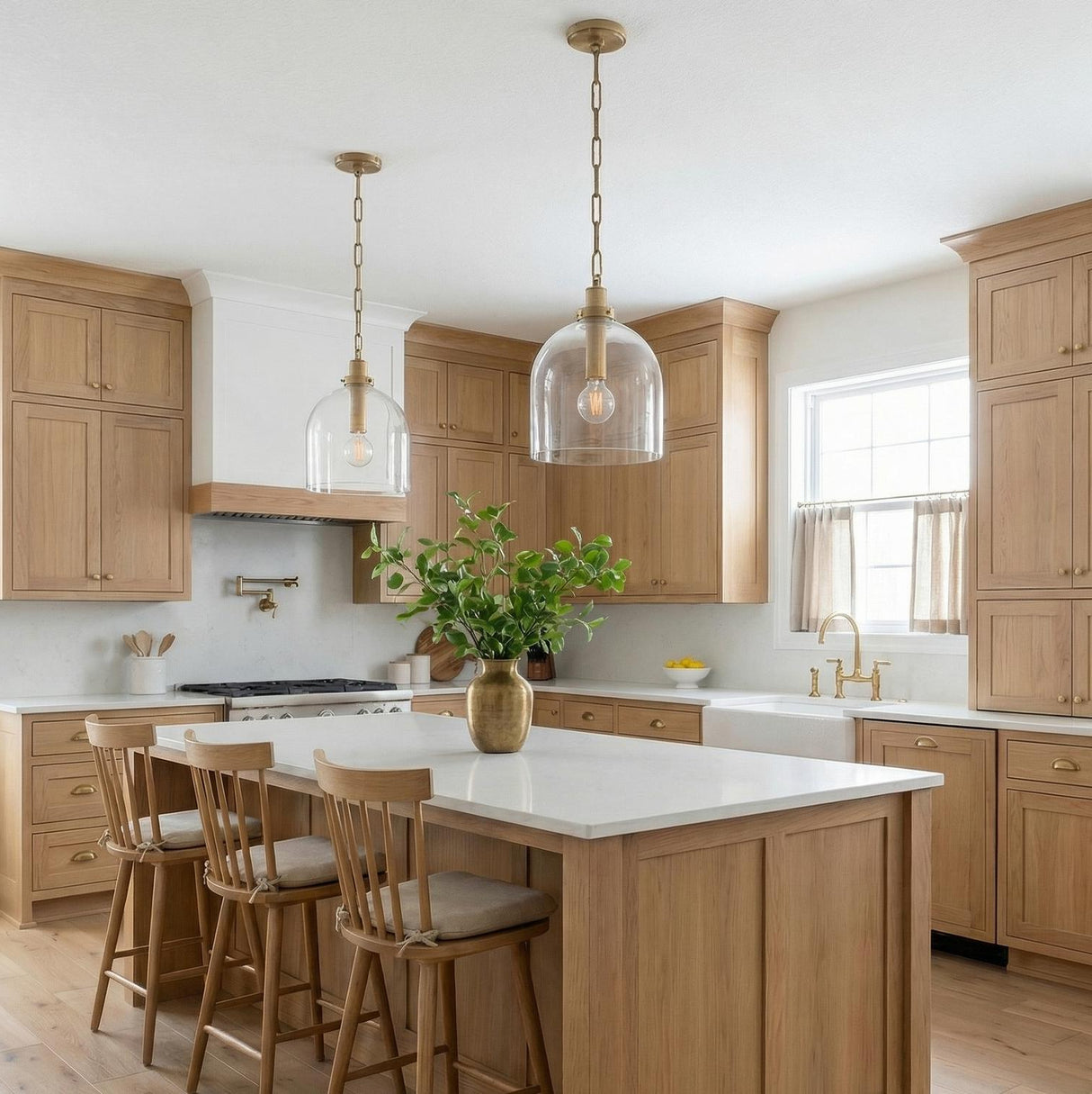 A farmhouse kitchen features two clear glass dome pendants with brushed brass accents over a white quartz island surrounded by light wood cabinetry and matching wooden stools.