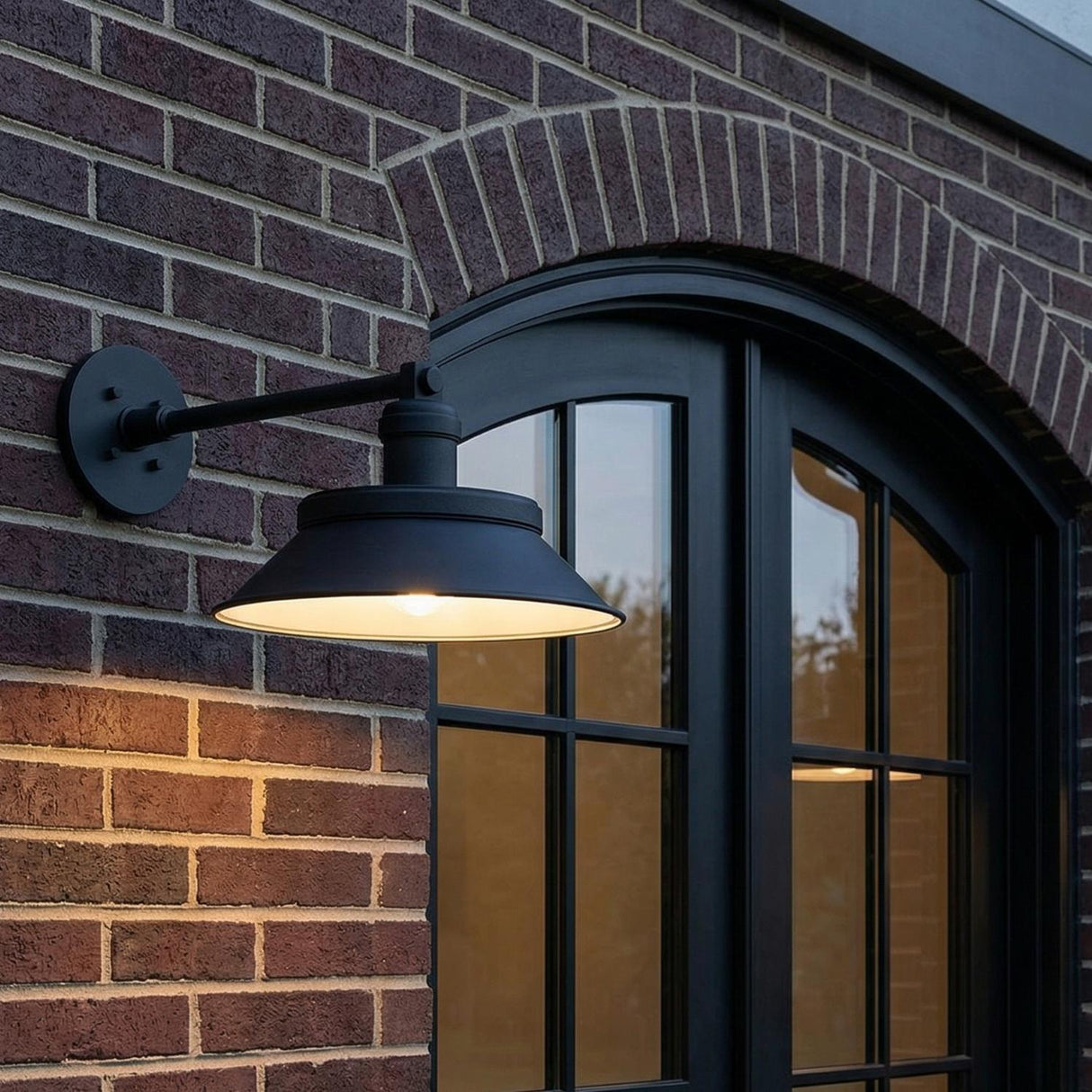 An outdoor entryway featuring the Vinya Outdoor Wall Light, Medium, Matte Black sconce mounted on a red brick wall beside an arched black window frame.