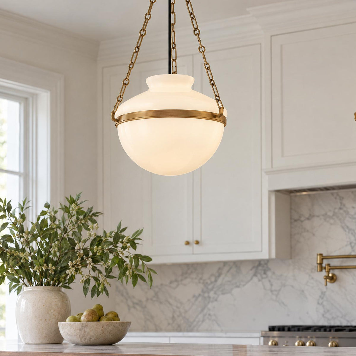 A transitional kitchen features the Capella Large Pendant in Patina Brass and Milk Glass hanging over a marble counter near white cabinets and a marble backsplash.