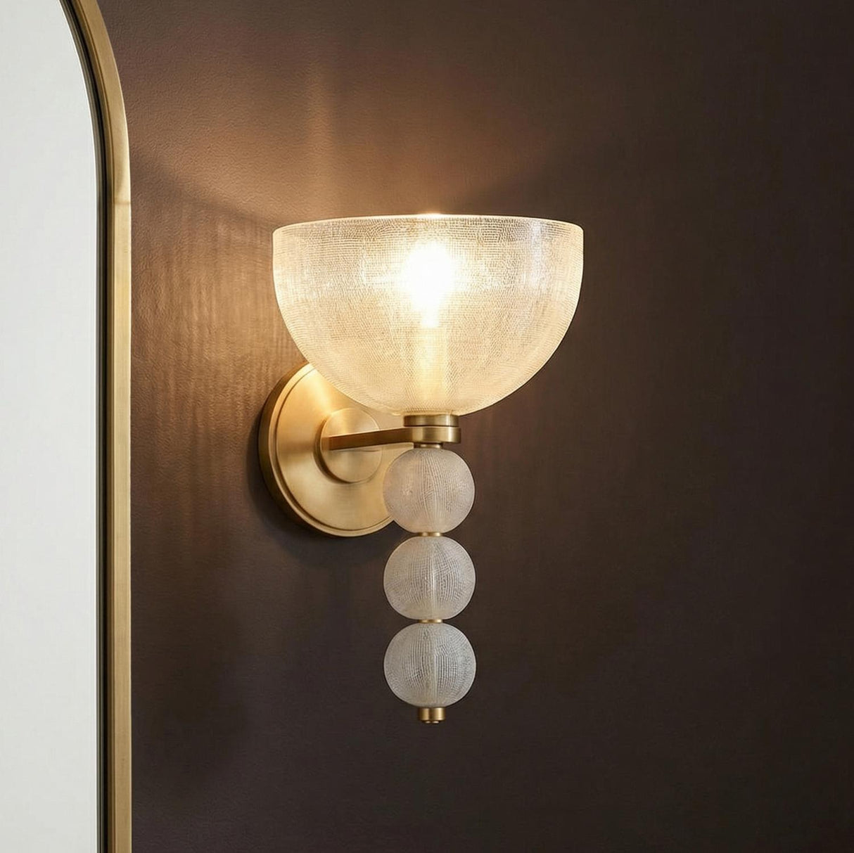 A contemporary bathroom featuring an Amity Wall Sconce in aged brass and textured glass installed on a dark brown wall next to a brass-framed mirror.