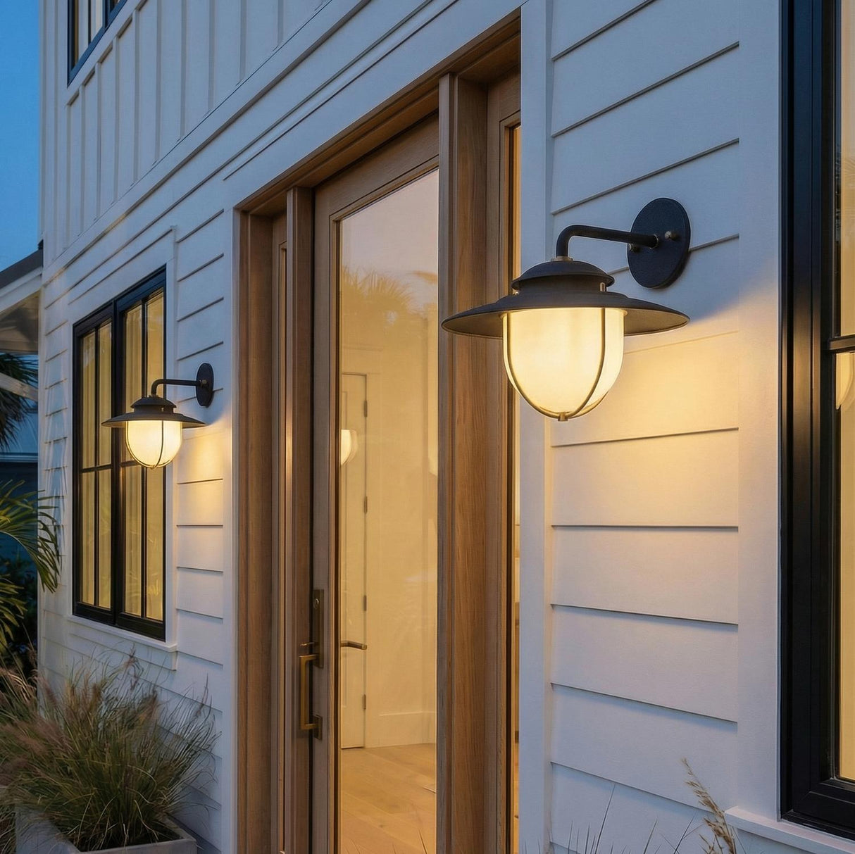 A modern farmhouse entryway featuring two forged iron and patina brass wall sconces with frosted glass globes mounted on white siding beside a wooden door.