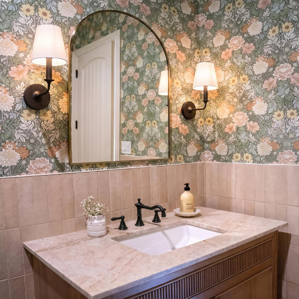 A cottage bathroom featuring two Harwick Wall Sconces in aged iron with white linen shades flanking an arched mirror above a marble vanity and floral wallpaper.