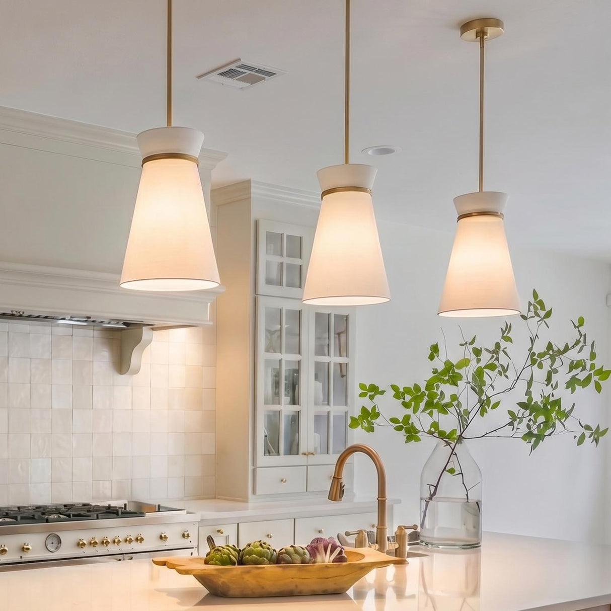 A modern farmhouse kitchen with three satin brass and white linen pendants hanging over a white island beside a brass faucet and wooden bowl.