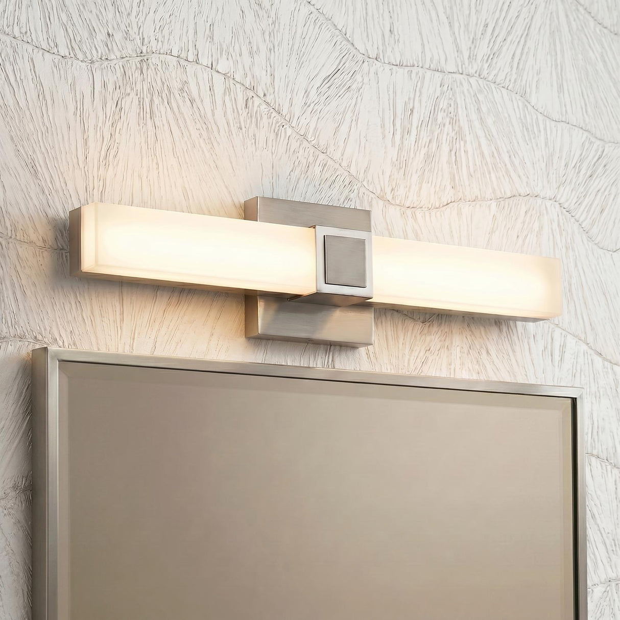 A modern bathroom features a Wendell LED Vanity Light 18", Brushed Nickel with frosted acrylic mounted over a framed mirror against wavy textured white wallpaper.