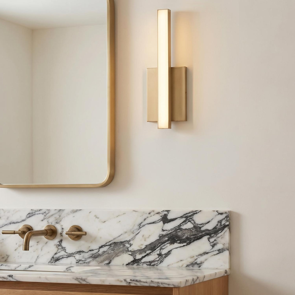 A contemporary bathroom with a Garrison LED Wall Sconce 13", Satin Brass mounted beside a gold mirror above a white marble backsplash and matching brass faucet.