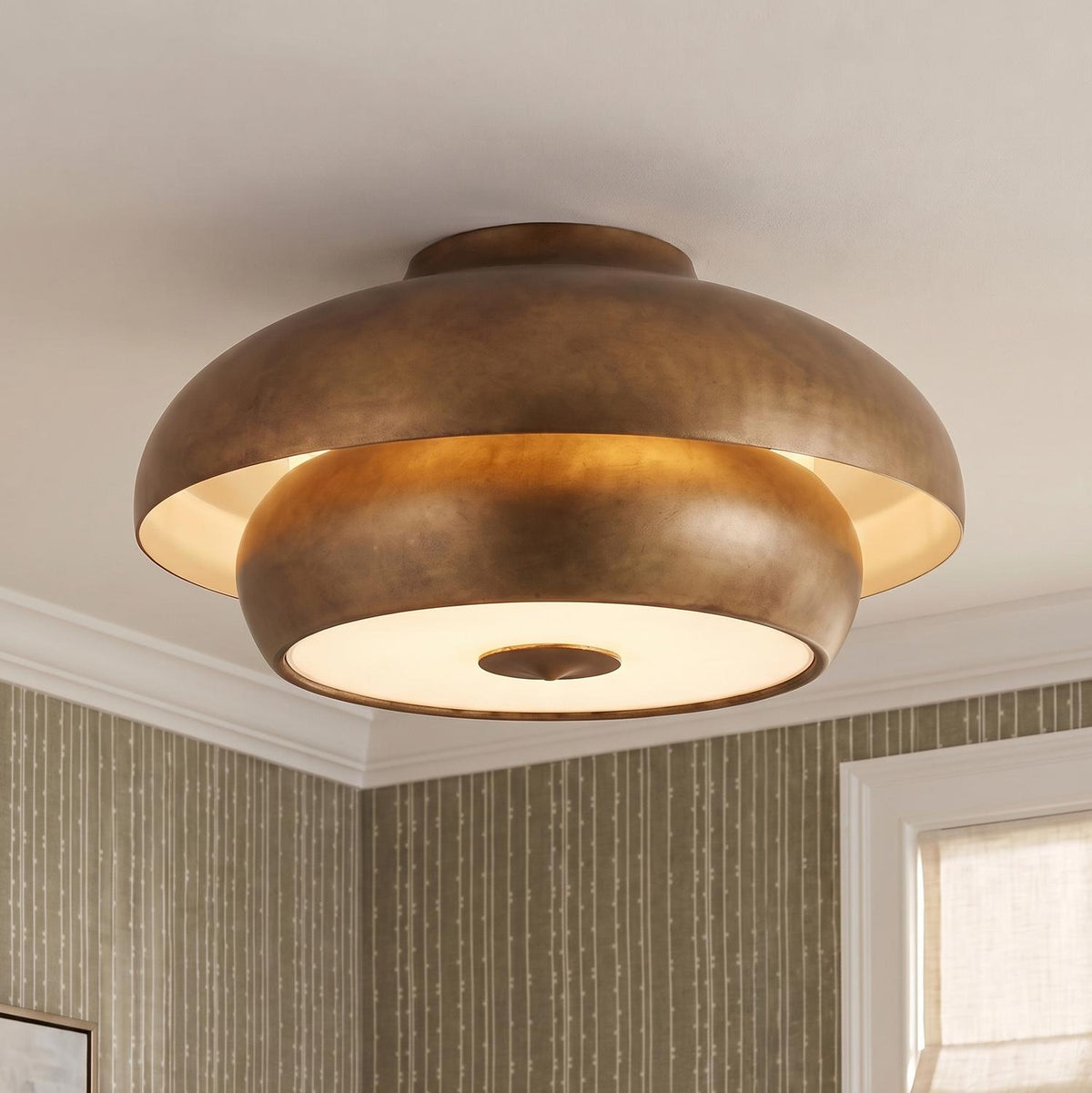 A transitional bedroom featuring a vintage gilded brass Garnett semi-flush mount on a standard ceiling above olive green striped wallpaper and a linen window shade.