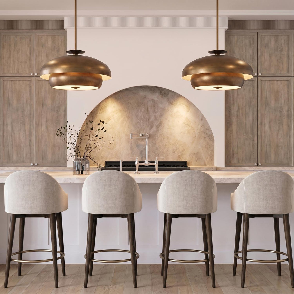 A transitional kitchen with two Garnett Large Pendants in vintage gilded brass over a white marble island, featuring four upholstered stools and an arched stone backsplash.