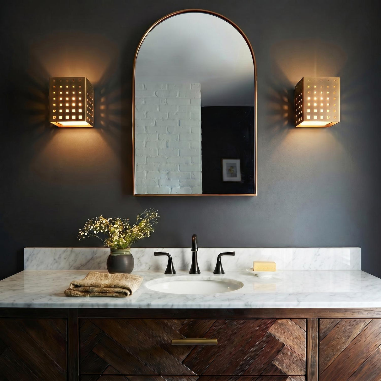 A contemporary bathroom featuring two Egan LED Wall Sconces in vintage brass flanking an arched mirror over a white marble countertop and chevron wood vanity.