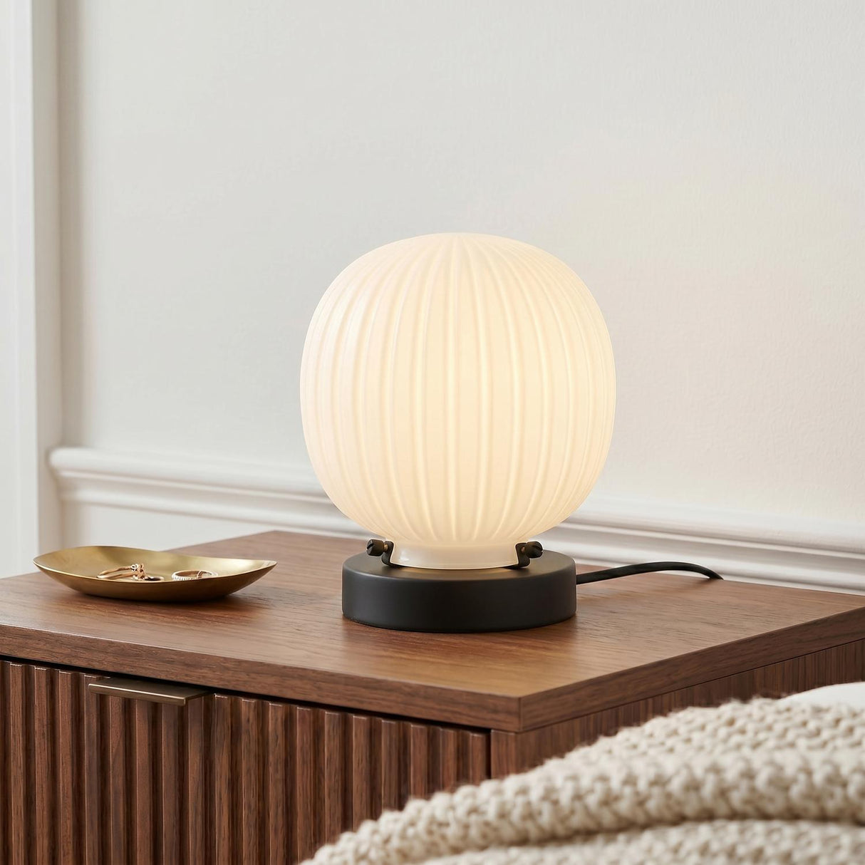 A mid-century bedroom featuring the Basile Small Table Lamp 8", Matte Black and Ribbed Milk Glass on a fluted walnut nightstand beside a cream knit blanket.
