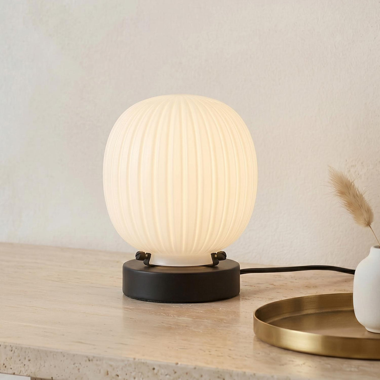 A modern living room featuring the Basile Small Table Lamp with matte black and ribbed milk glass on a travertine console table beside a round brass tray.