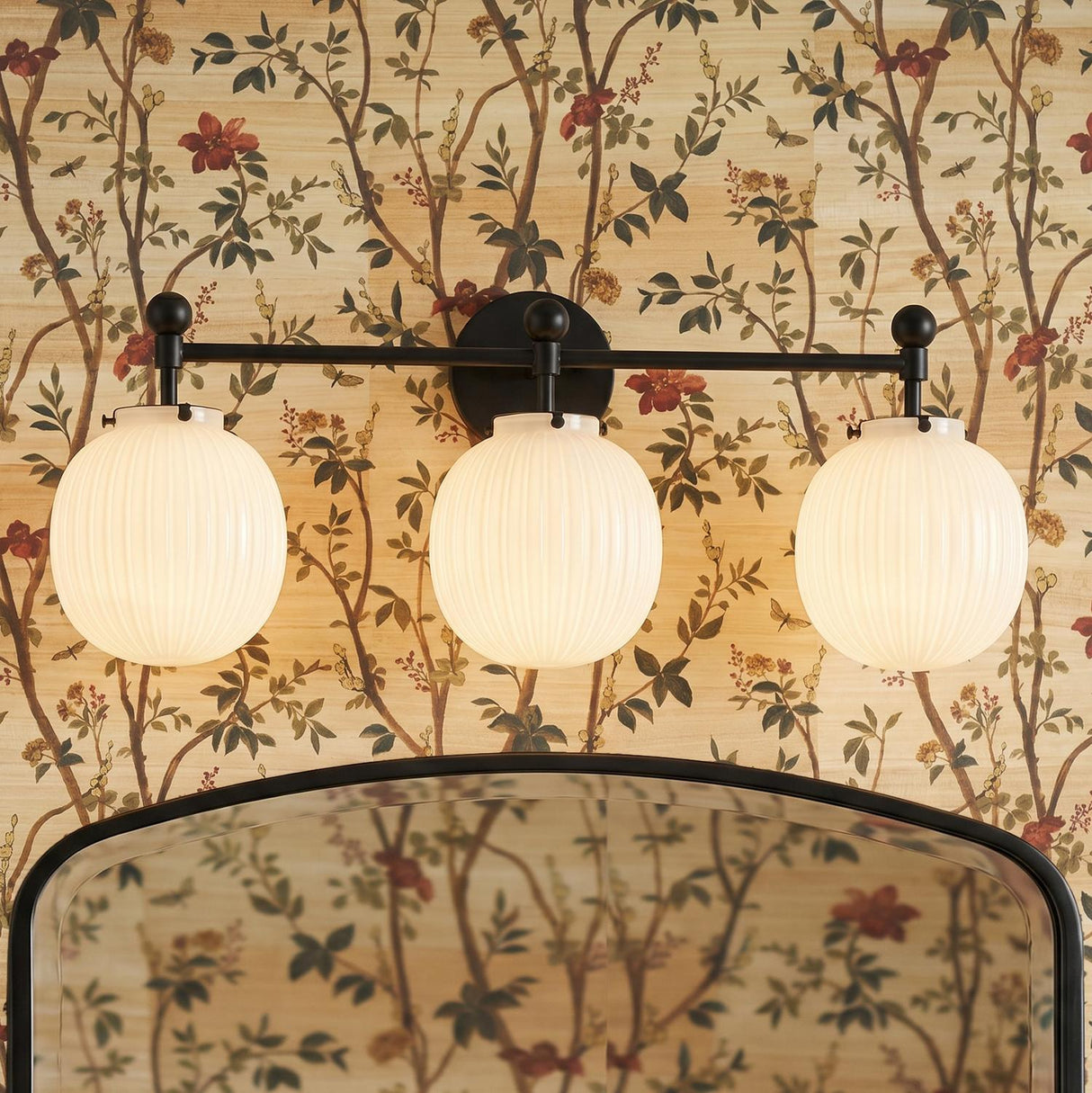 A traditional bathroom with the Basile 3 Light Vanity, Matte Black and Ribbed Milk Glass installed above a black arched mirror on botanical wallpaper.