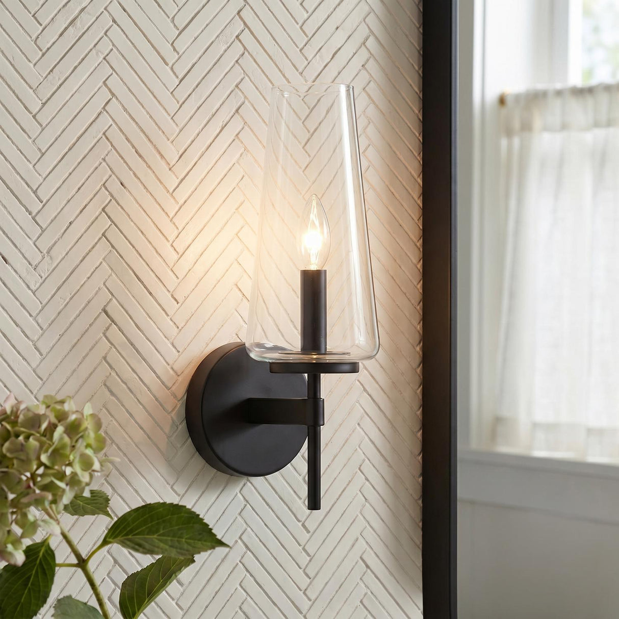A transitional bathroom with the Darlene Wall Sconce in matte black featuring a clear tapered glass shade mounted on a white herringbone tile wall beside a mirror.