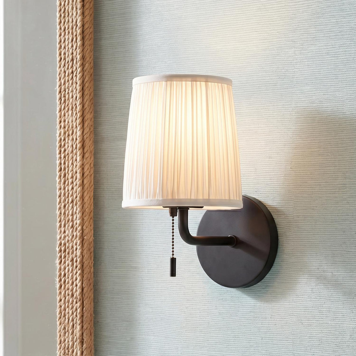 A coastal foyer with a matte black Daina Wall Sconce and white pleated linen shade mounted beside a rope framed mirror on light blue textured wallpaper.