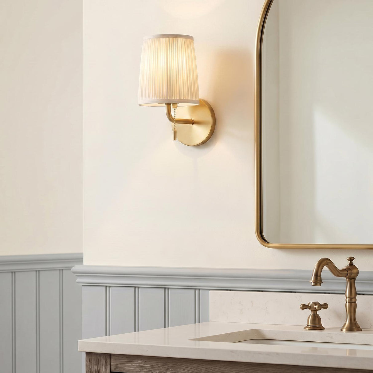 A transitional bathroom with a brushed brass wall sconce featuring a white pleated fabric shade mounted beside a gold-framed mirror above a marble vanity and gray wainscoting.