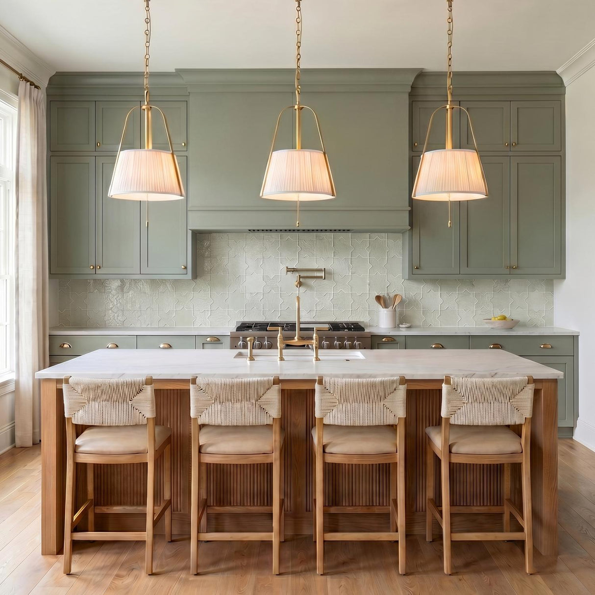 A transitional kitchen with three brushed brass pendants and white linen shades suspended over a white marble island and sage green cabinetry with tile backsplash.