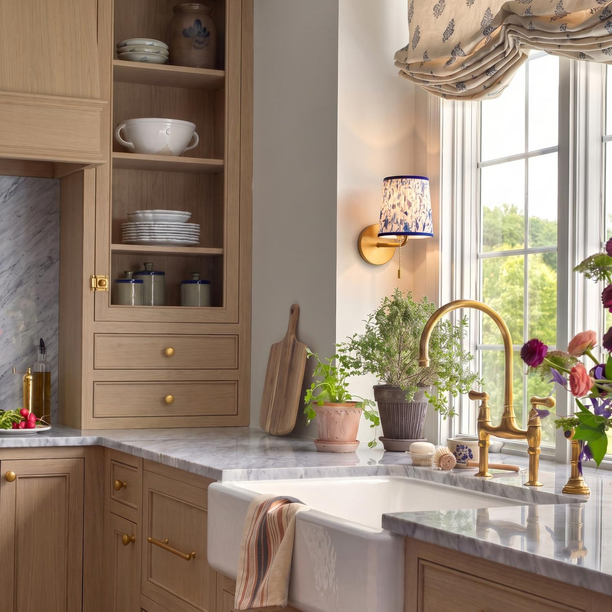 A traditional kitchen with the Daina Wall Sconce in brushed brass and blue floral mounted beside a window over a marble countertop and apron sink.