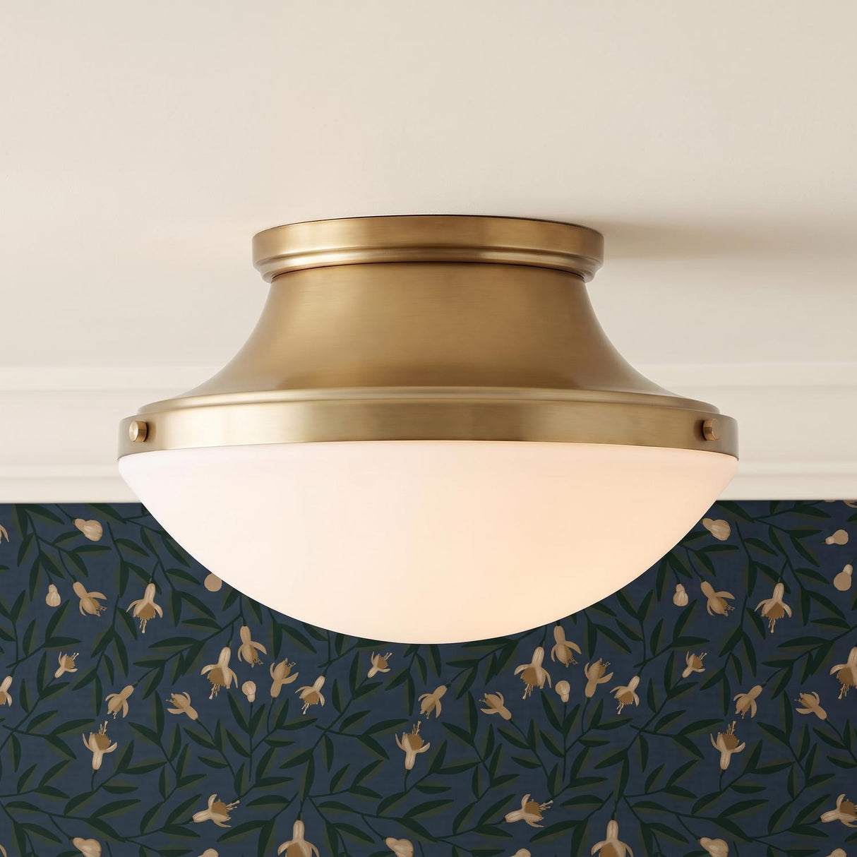 A transitional foyer with the Georgia Flush Mount in an aged brass finish and opal glass shade mounted to a standard ceiling above navy floral wallpaper.