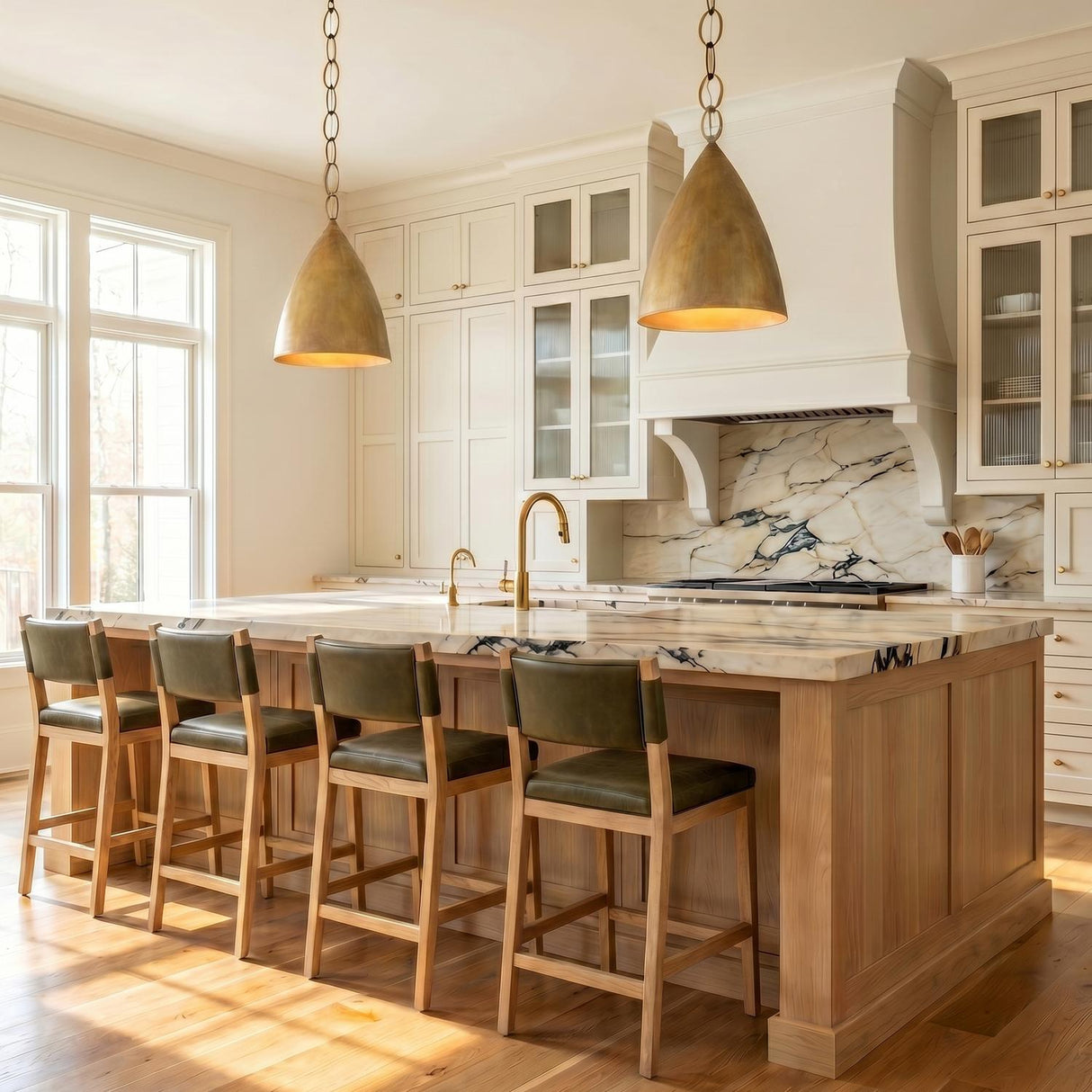 A transitional kitchen with two feathered brass cone pendants hanging over a marble topped wood island featuring cream shaker cabinets and a matching marble backsplash.