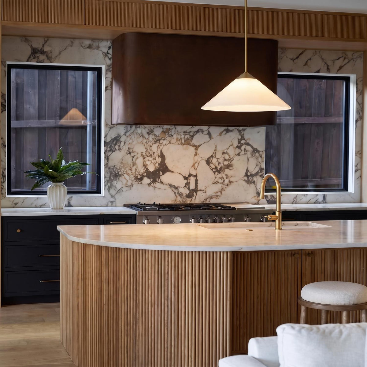 A contemporary kitchen with a matte brass and opal glass cone pendant over a curved fluted wood island with a marble countertop and brass faucet.