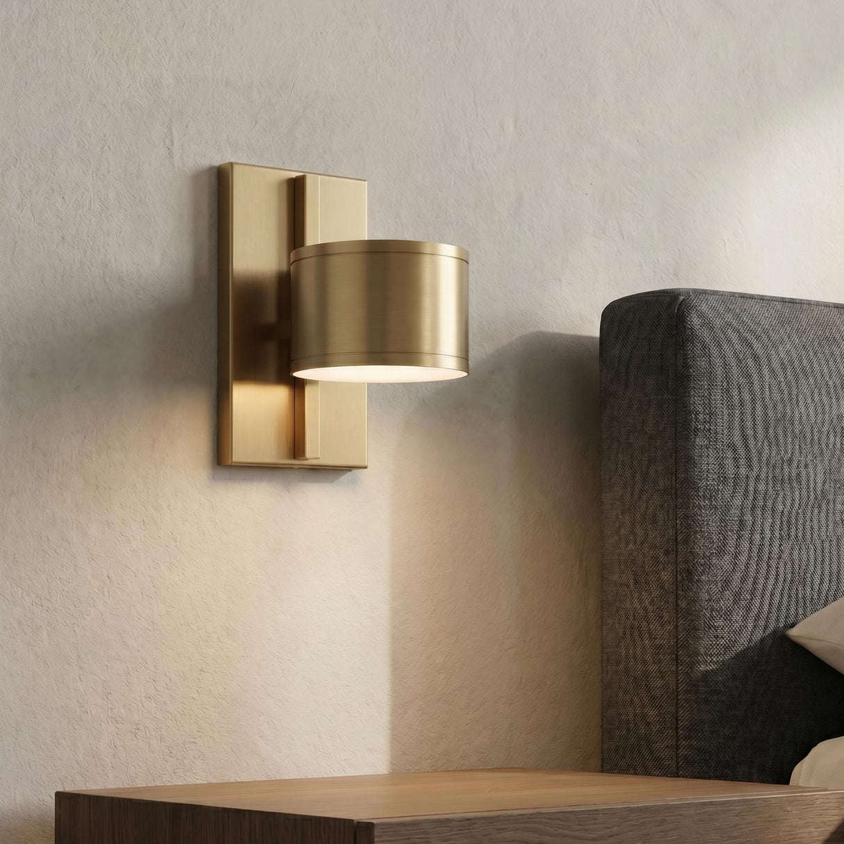 A modern bedroom with an aged brass Carmine LED Wall Sconce mounted on a textured beige wall beside a gray upholstered headboard and wood nightstand.