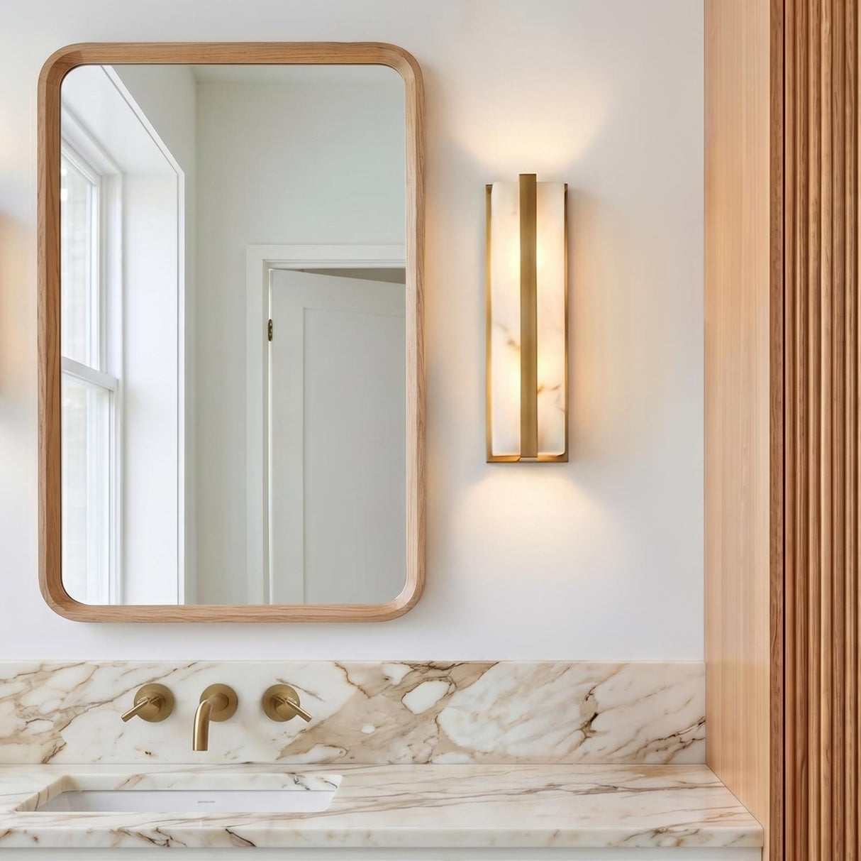 A modern bathroom with an Azura LED Wall Sconce 16", Aged Brass and Alabaster mounted beside a rounded oak mirror above a marble vanity counter.