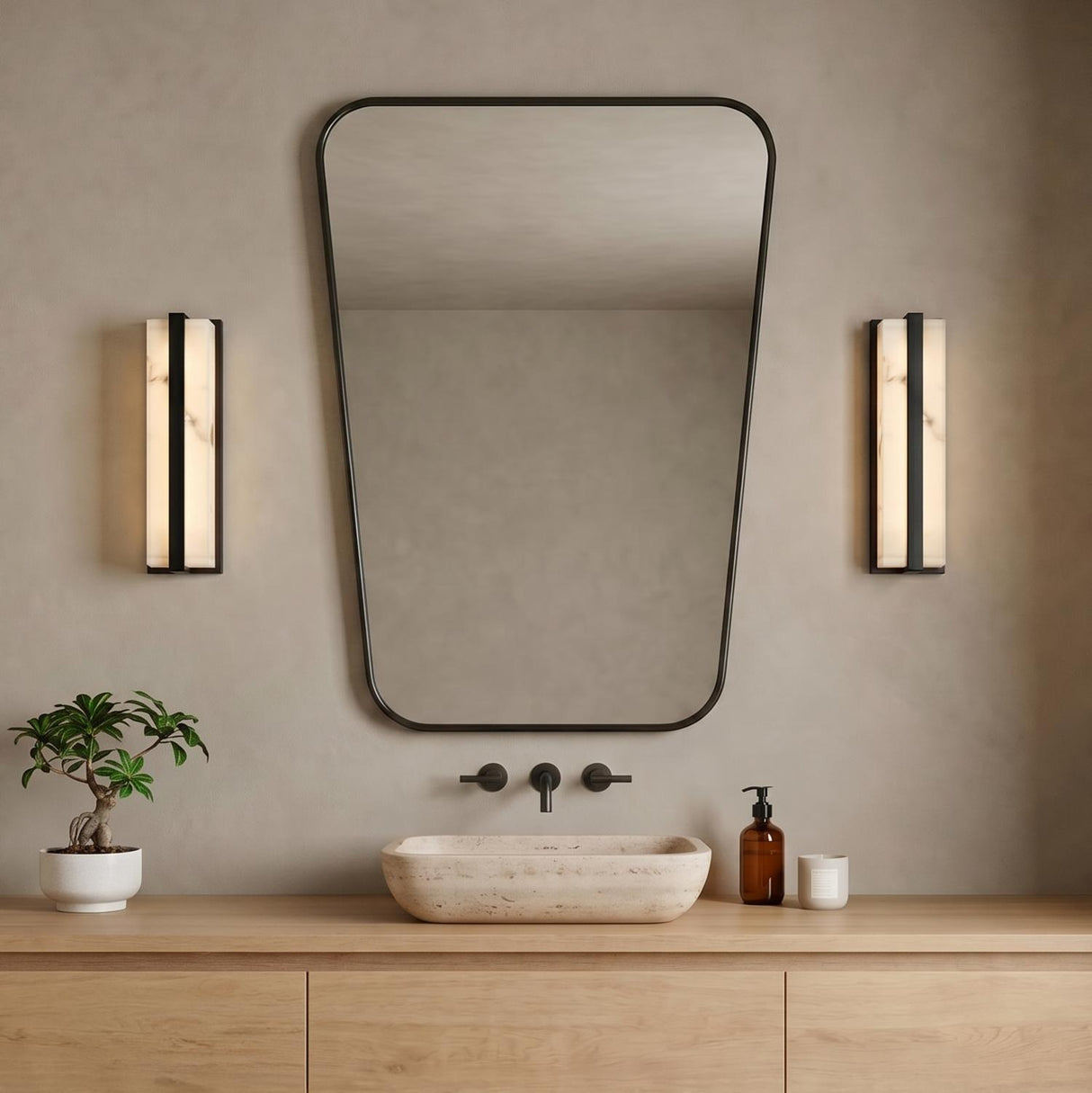A minimalist bathroom with two matte black and alabaster wall sconces flanking a rounded rectangular mirror above a light wood vanity and stone vessel sink.