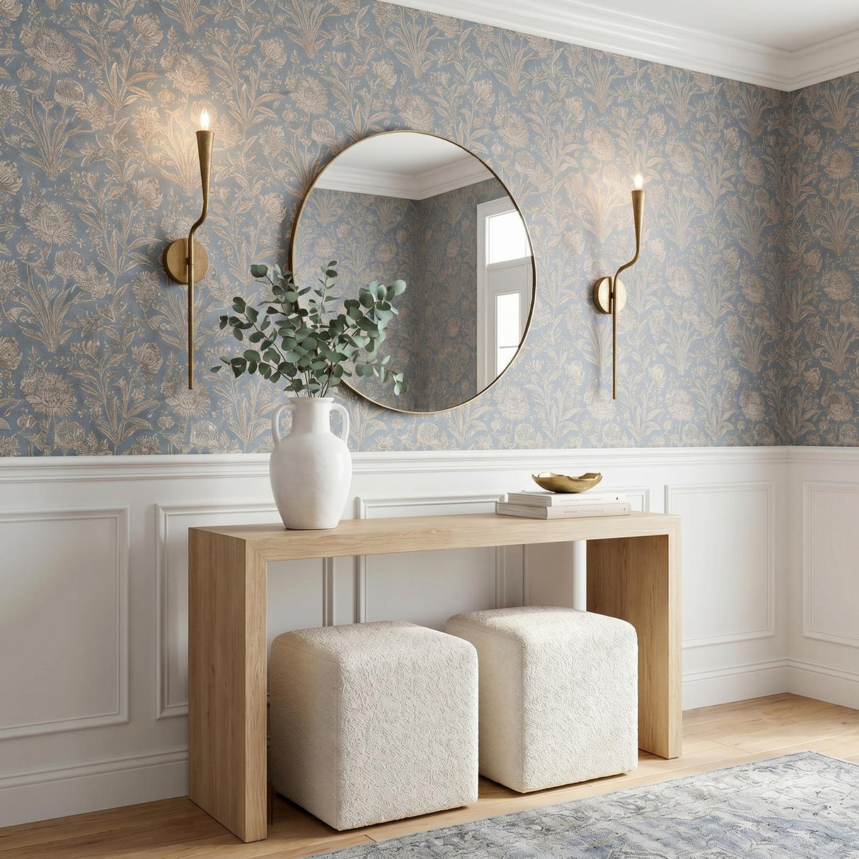 A transitional foyer with two Myra Wall Sconces in artisan brass flanking a round gold mirror above a light wood console table and blue floral wallpaper.