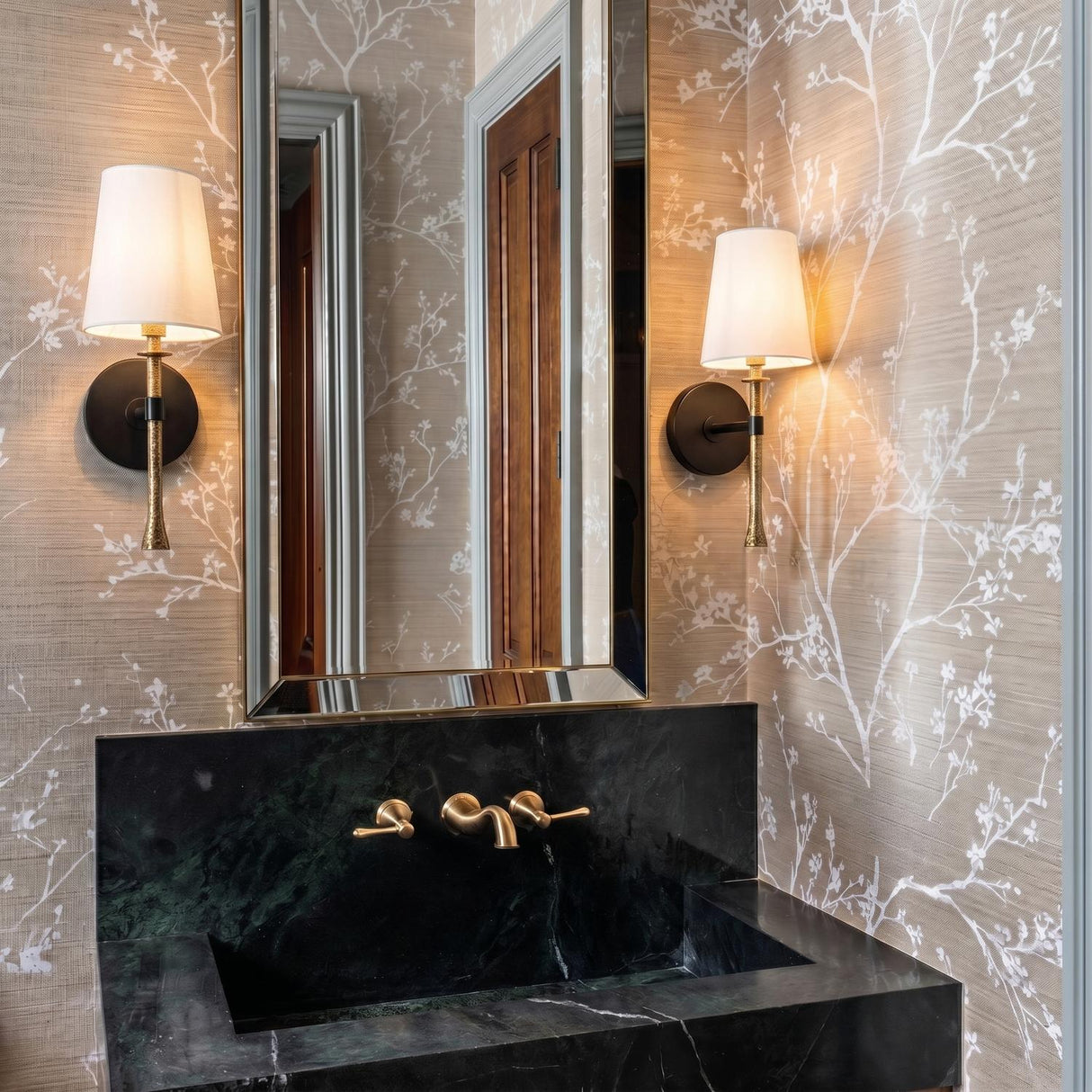 A transitional bathroom with two matte black and hammered brass Gerald Wall Sconces flanking a tall mirror above a dark green marble sink and botanical wallpaper.