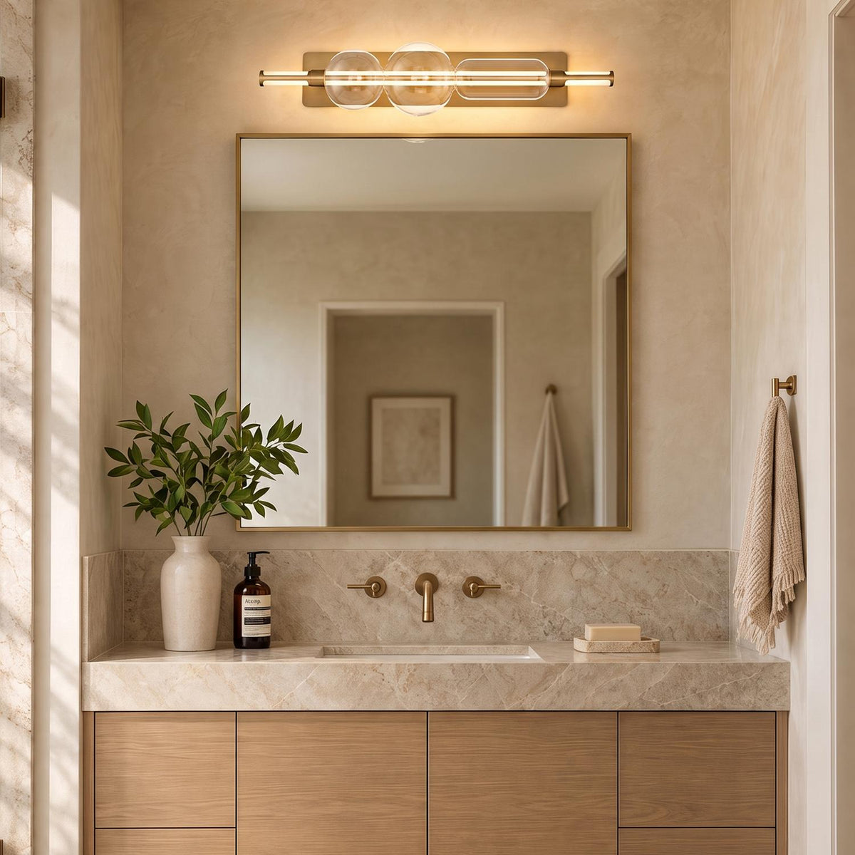 A modern bathroom features the Latimer LED Vanity Light 34", Brass and Clear Glass mounted above a brass mirror, marble countertop, and light wood cabinet.