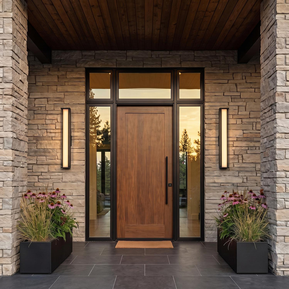 A modern home entry featuring two matte black Siran Outdoor LED Wall Sconces flanking a walnut front door on a stone facade with purple flowers in black planters.