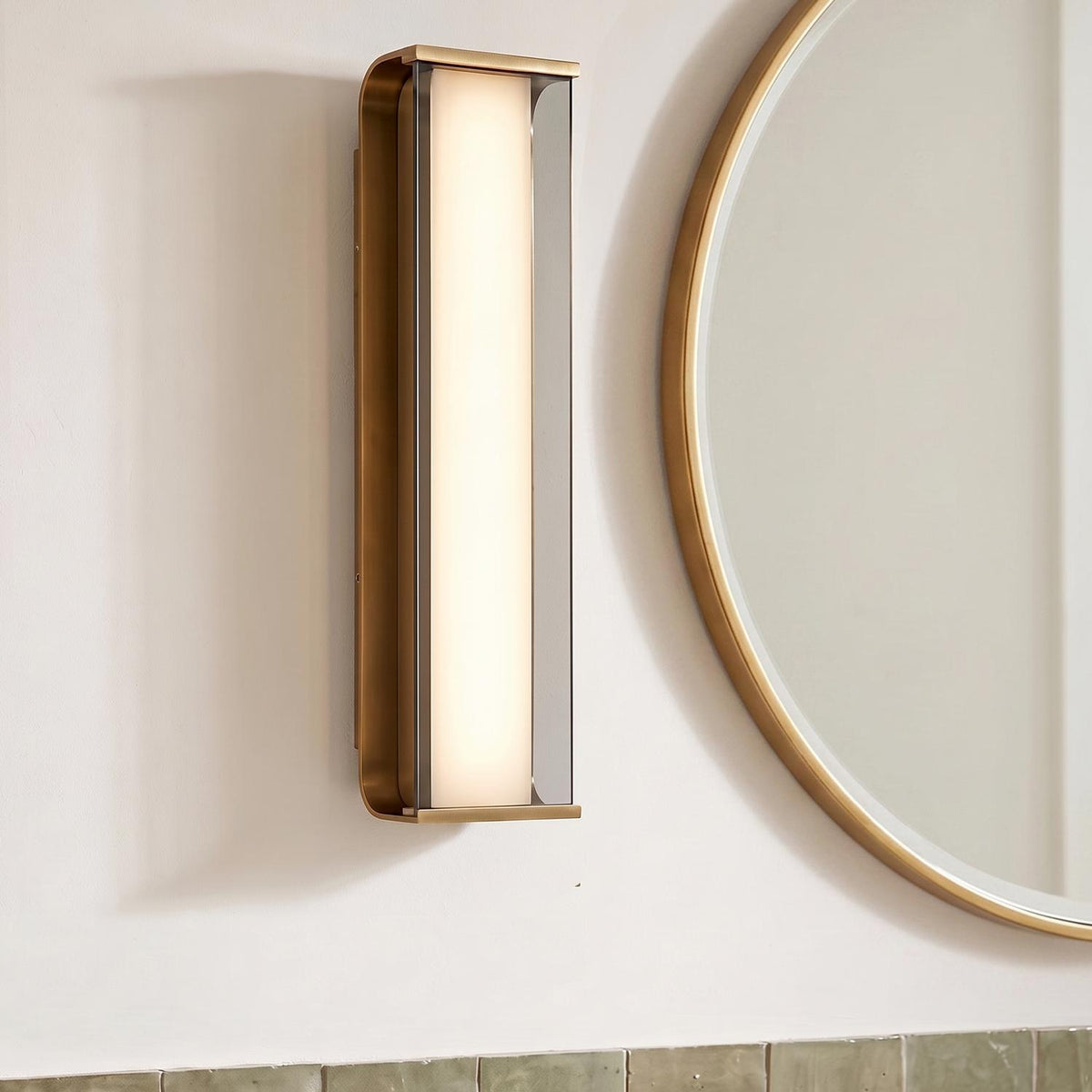 A contemporary bathroom with a Siran Large LED Wall Sconce in antique brass and smoke glass mounted beside a circular gold mirror above green tiling.