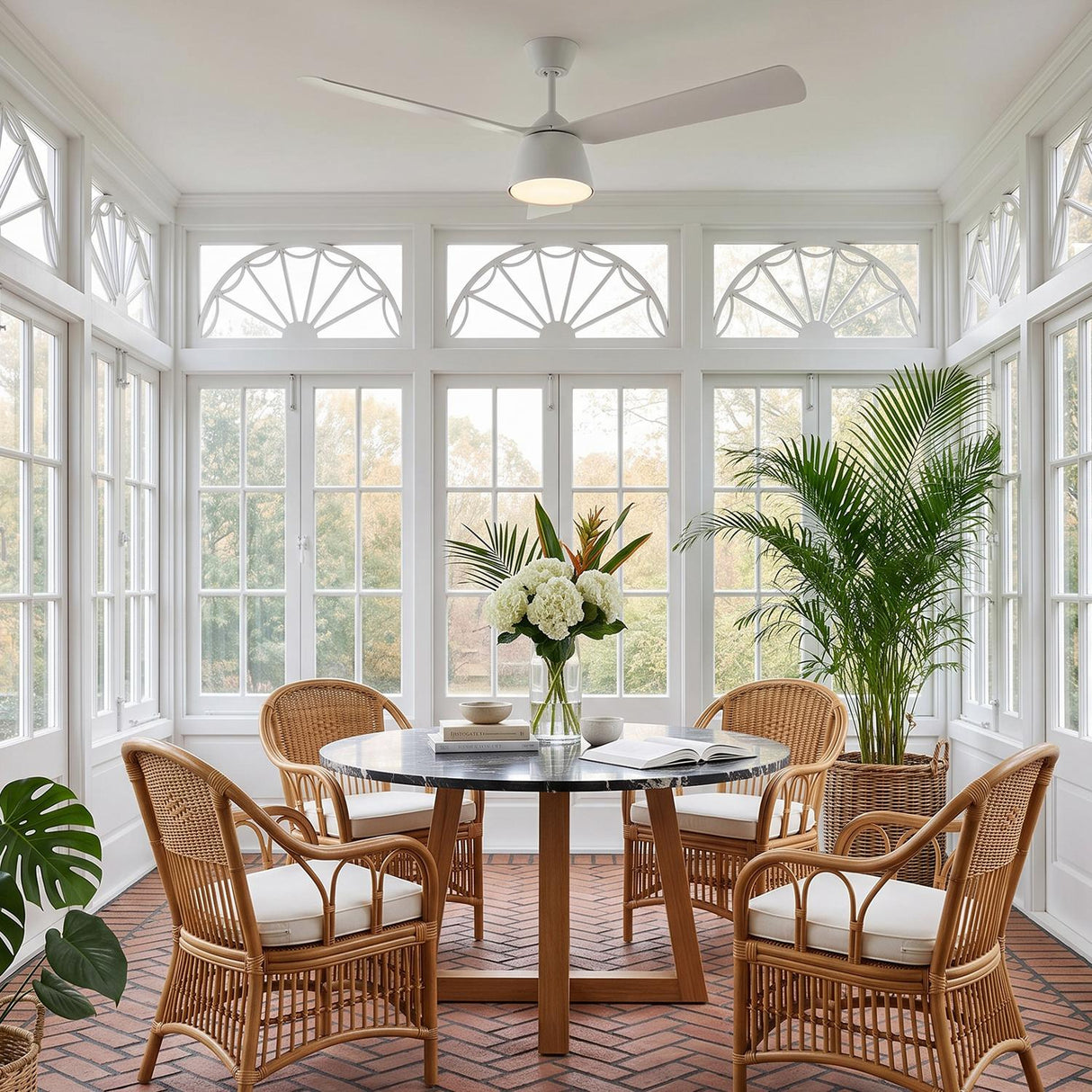 A coastal dining room featuring the Linnet LED Ceiling Fan 60-inch, Matte White over a black marble table and wicker chairs surrounded by white windows and brick flooring.