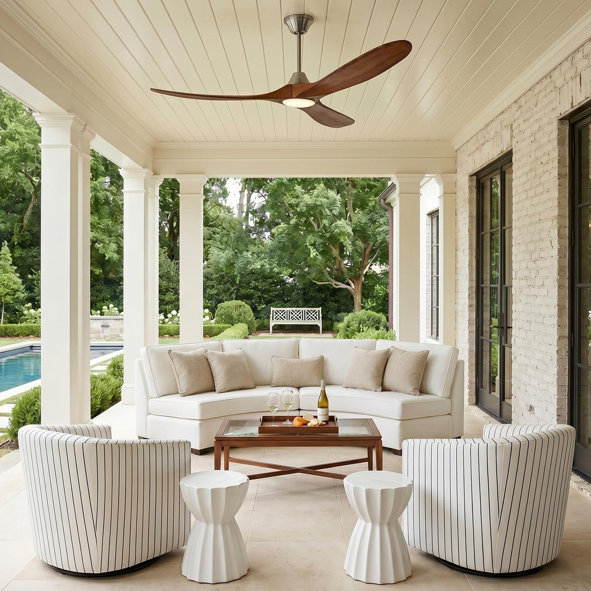 A modern farmhouse porch featuring the Egret LED Ceiling Fan 60" in koa wood and brushed steel mounted above a curved white sectional and white brick walls.