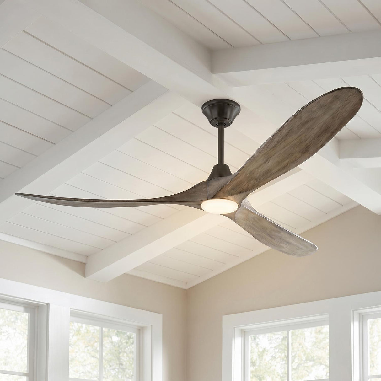 A modern farmhouse living room with the Egret LED Ceiling Fan in gray wash oak and aged pewter finish on a white beamed ceiling above bright windows.