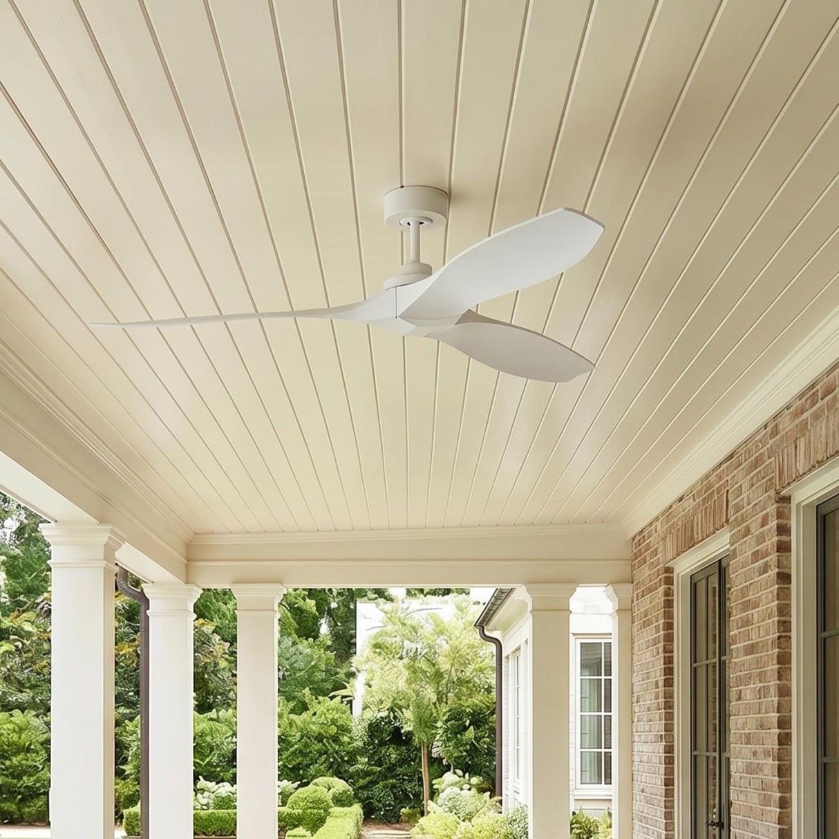 A traditional home porch with the Heron Outdoor Smart Ceiling Fan 60", Matte White on a shiplap ceiling beside a red brick wall, white columns, and boxwood hedges.