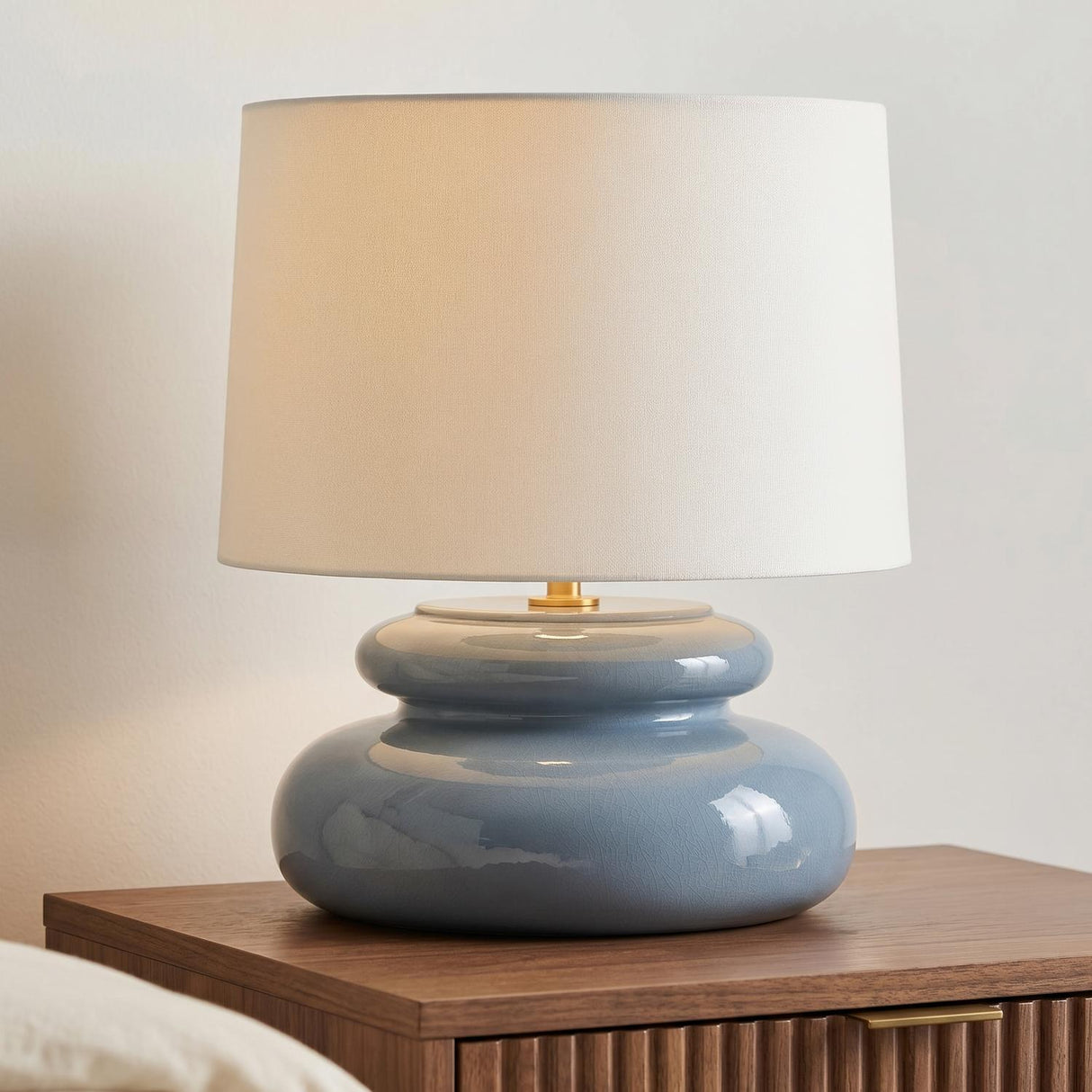 A modern bedroom featuring the Ellsworth Table Lamp 20", Blue Crackle Ceramic with a white linen shade on a fluted walnut nightstand beside a white wall.