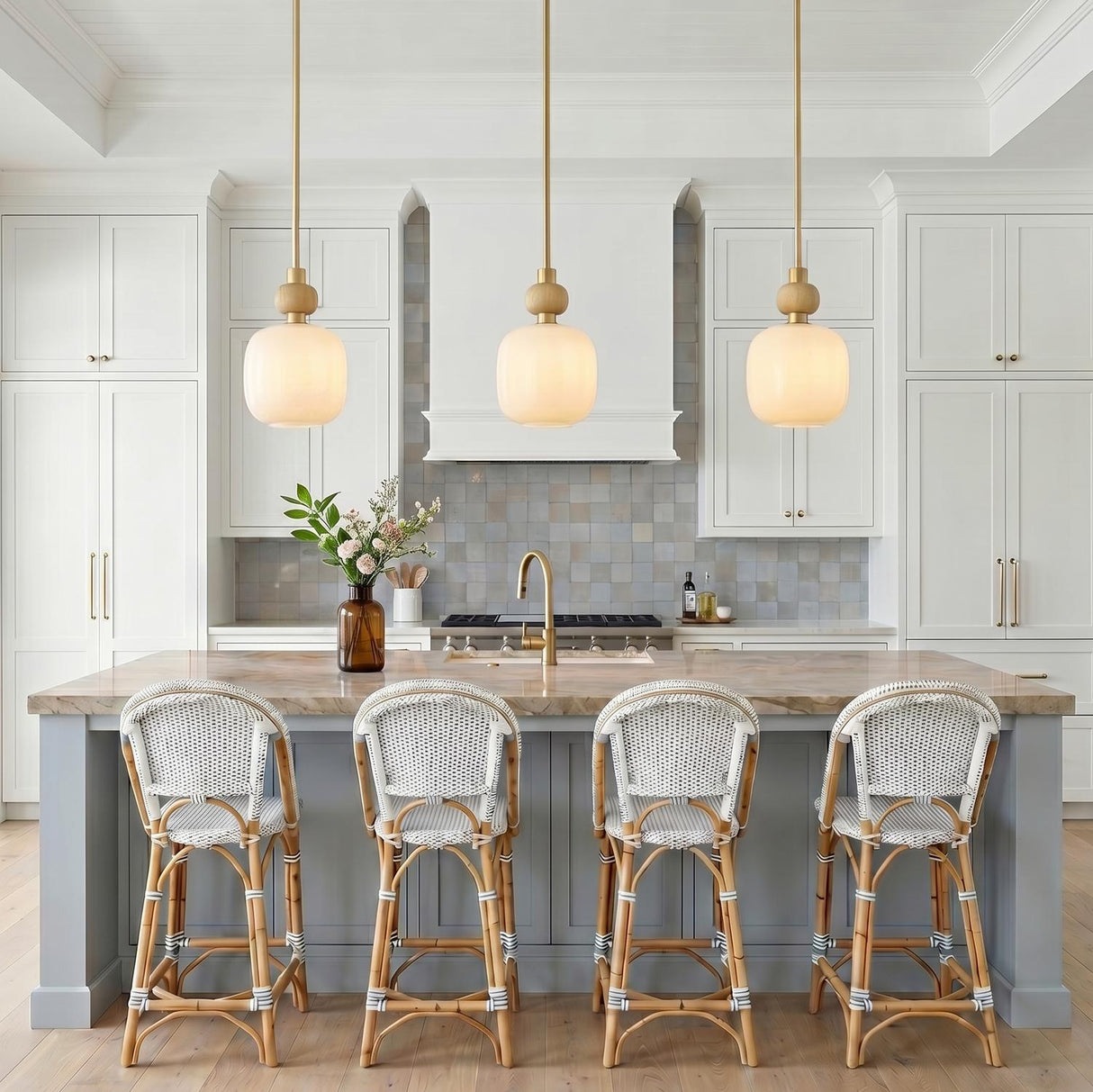 A coastal kitchen with three Norden Small Pendants in aged brass and milk glass over a stone-topped island with woven rattan stools and coffered ceilings.