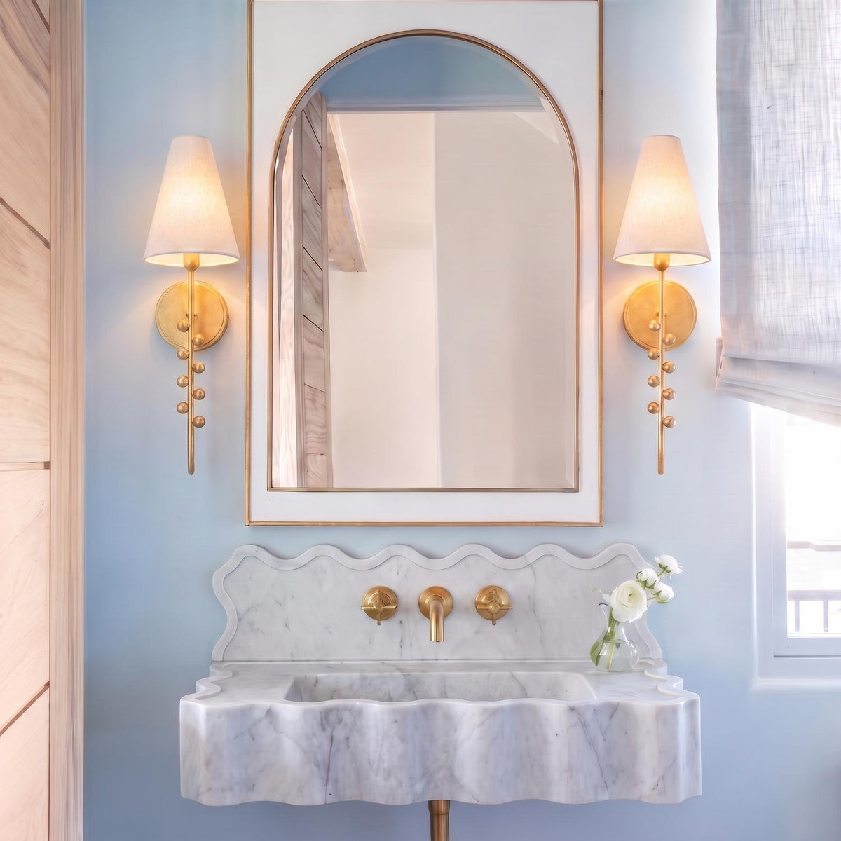 A transitional bathroom features two Linnea Wall Sconce 21", Gold Leaf fixtures with fabric shades flanking an arched mirror above a scalloped marble vanity.