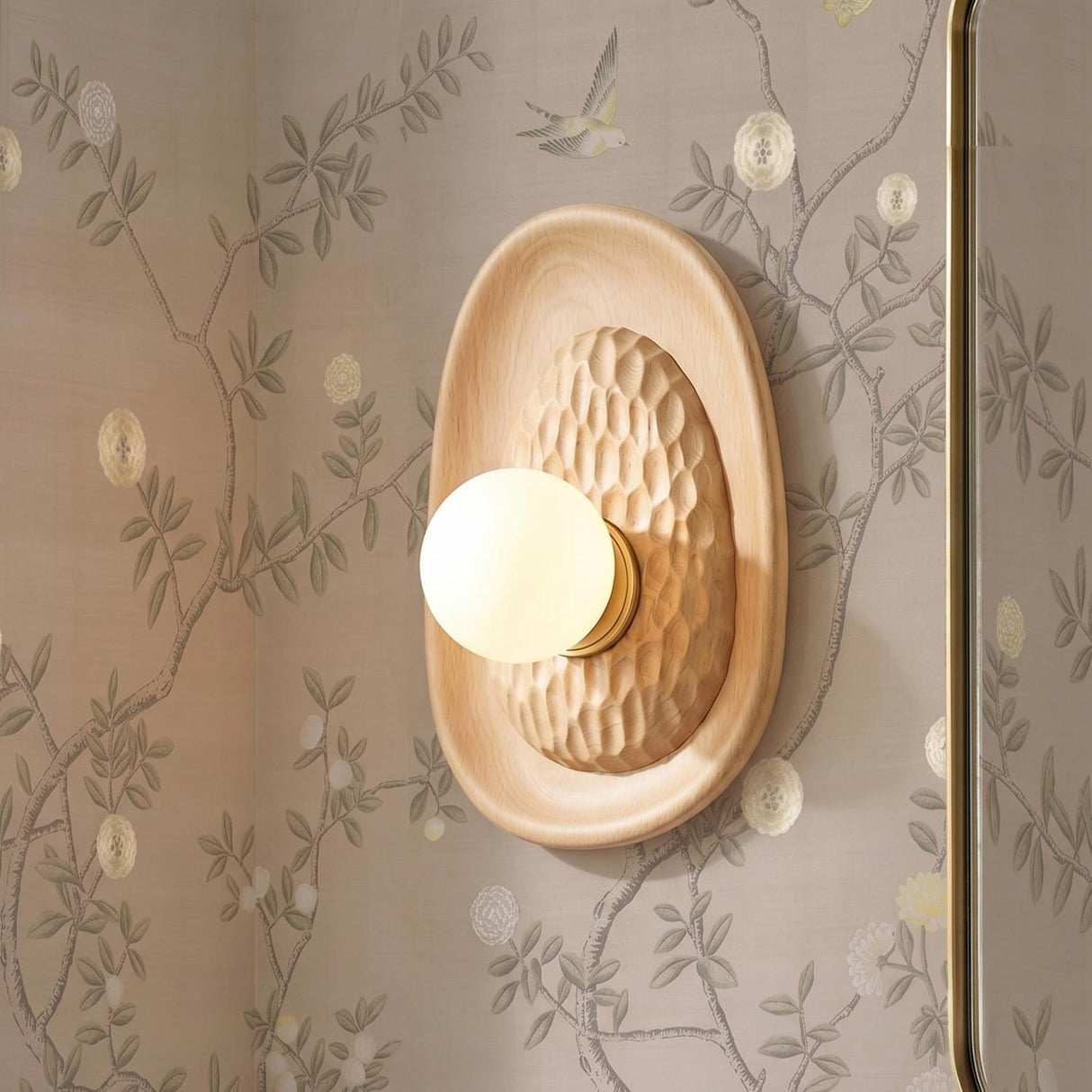 A transitional bathroom with a Sybella Wall Sconce in beech wood and aged brass featuring a hand-carved texture and globe bulb on floral wallpaper beside a mirror.