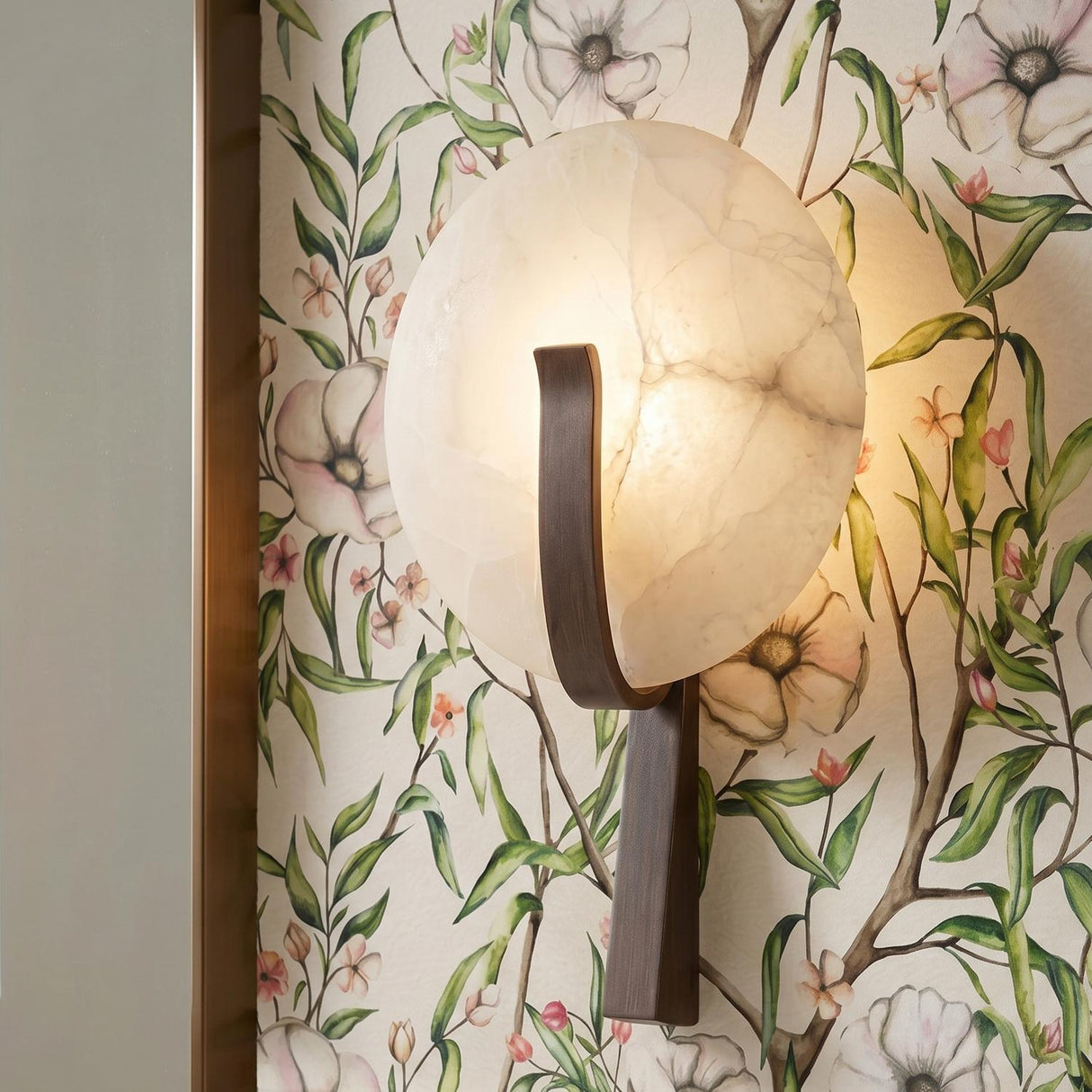 A transitional foyer with the Zahara LED Wall Sconce 16", Alabaster and Bronze Leaf mounted against a floral wallpaper featuring green leaves and cream-colored blossoms.