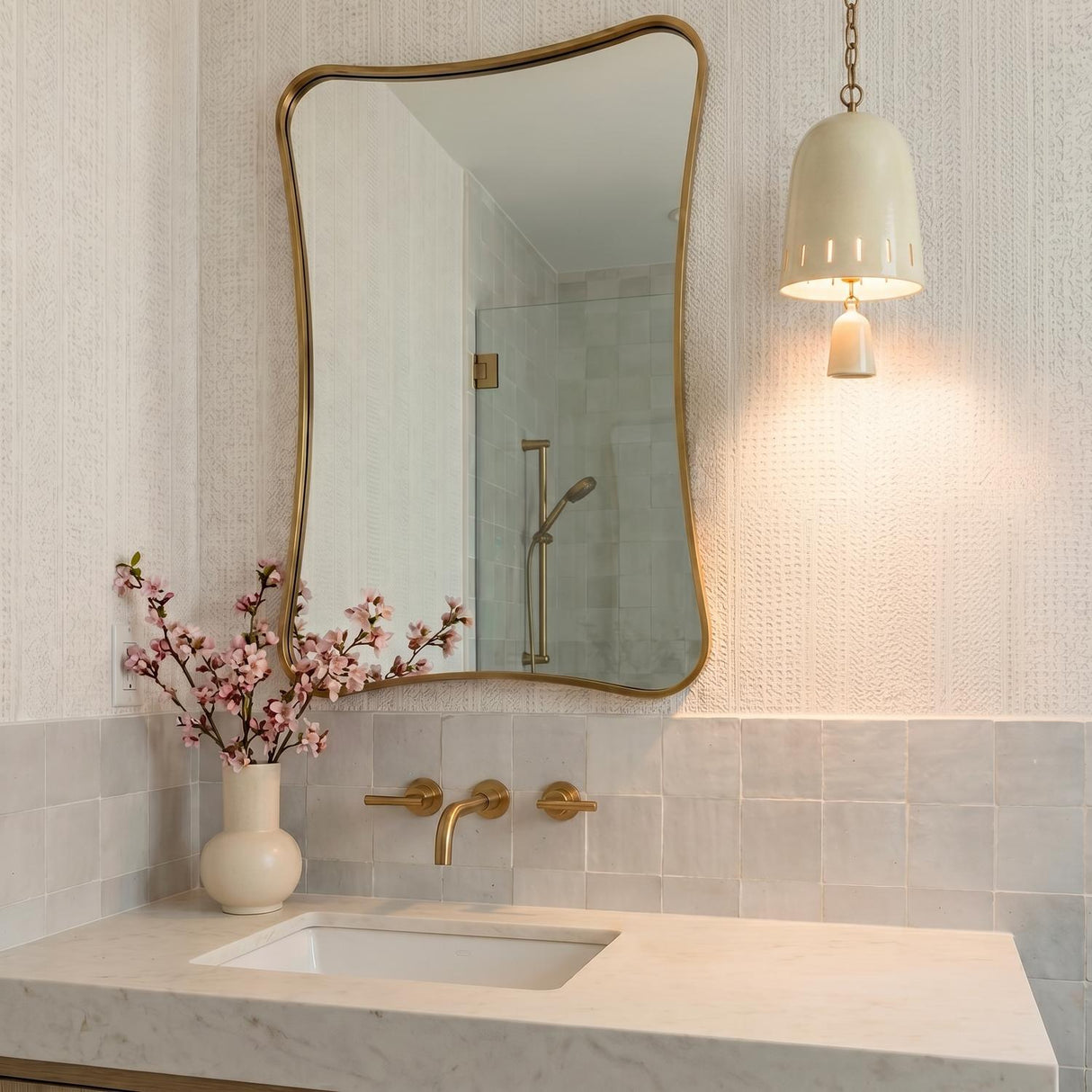 A mid-century bathroom featuring an ivory glaze and patina brass Briony Small Pendant hanging beside a wavy gold mirror above a white marble vanity.