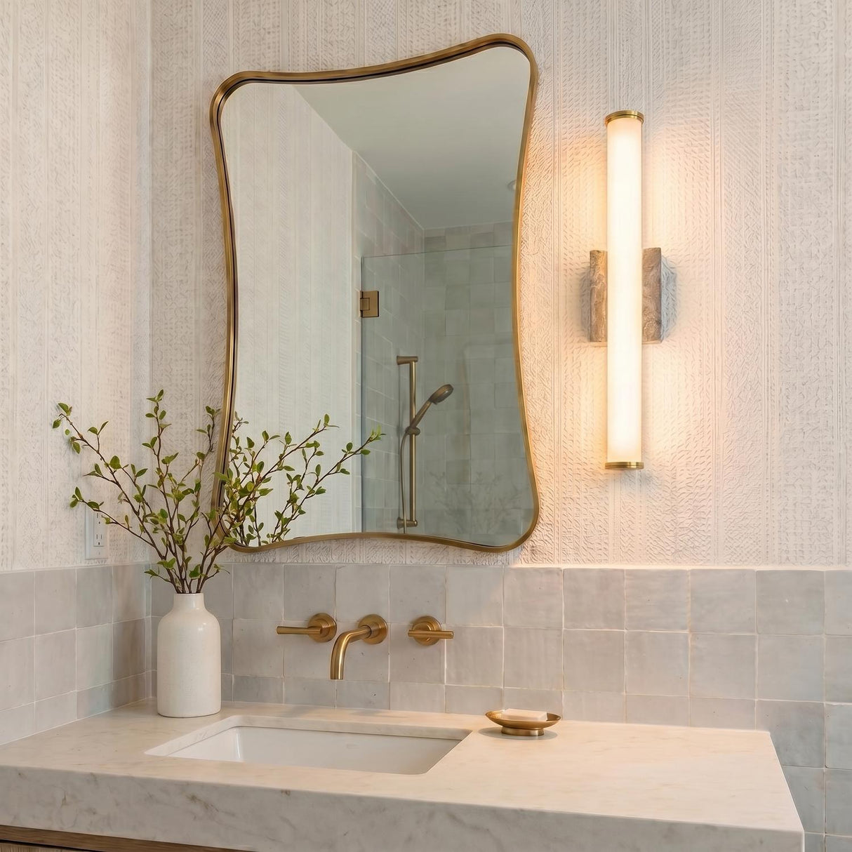 A contemporary bathroom with a Gaven LED Wall Sconce in gray travertine and aged brass mounted beside an organic mirror above a white marble vanity with brass faucets.
