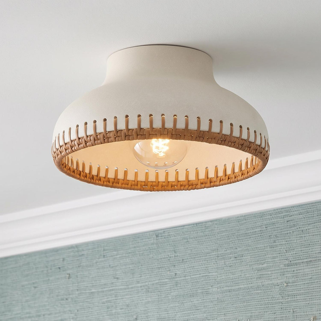 A coastal bedroom featuring the Nadira semi-flush mount in cream ceramic and rattan with aged brass accents above blue grasscloth wallpaper and white molding.