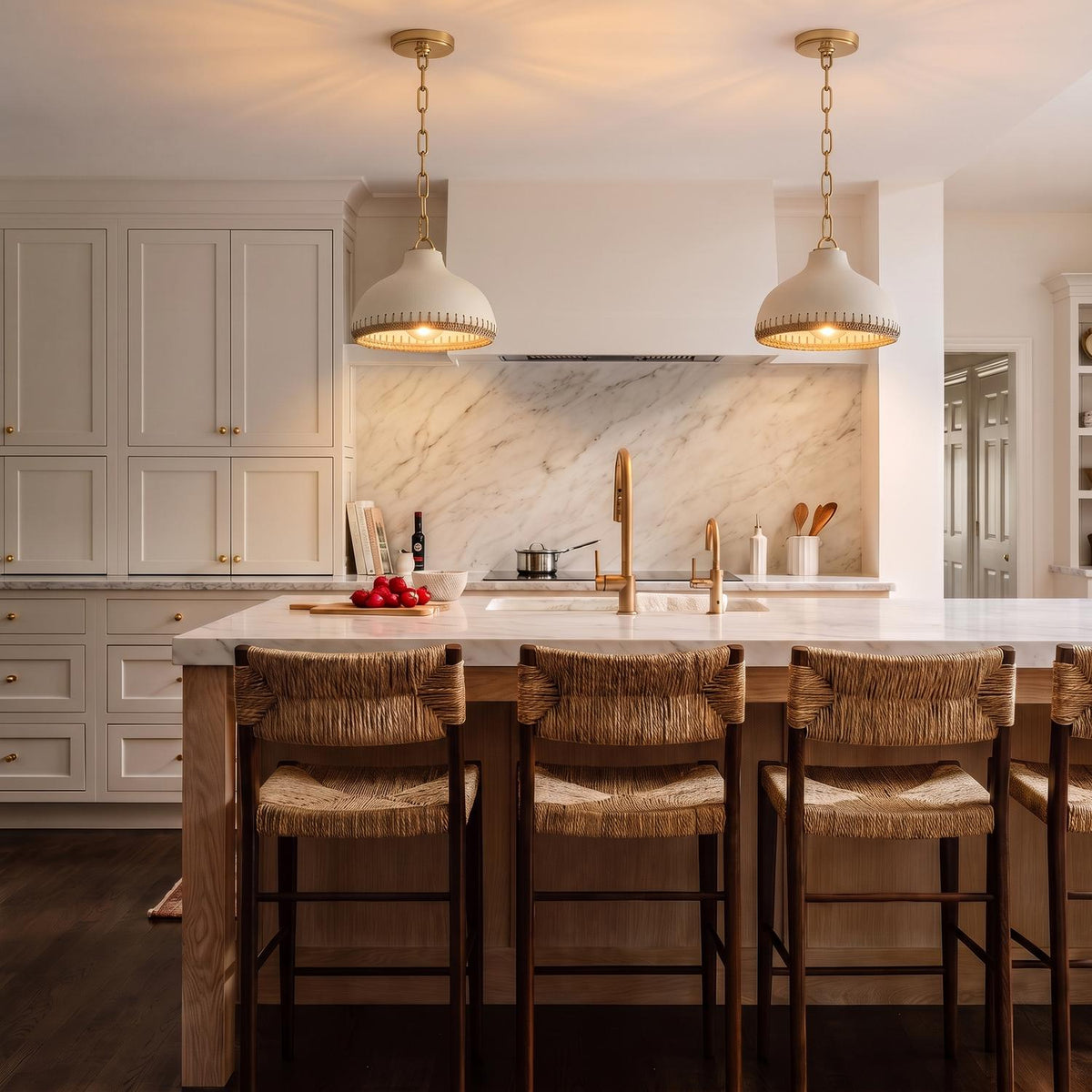 A transitional kitchen with two cream and rattan pendants featuring aged brass hardware over a white marble island with woven stools and white shaker cabinets.